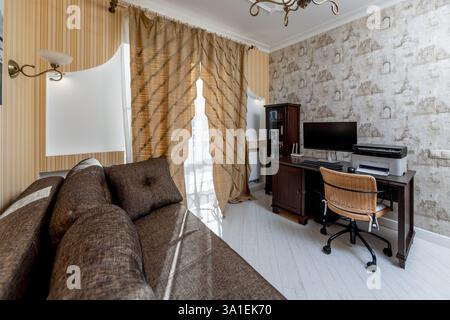 Ein Wohnzimmer mit einer braunen Couch und einem braunen Stuhl mit einem Computertisch in der Ecke. Das Zimmer hat eine moderne und komfortable Atmosphäre Stockfoto