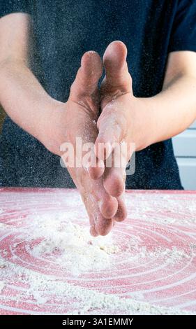 Die Hände eines Kindes klatschen mit Mehl über den Tisch mit rotem Backblech darauf Stockfoto