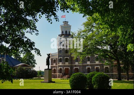 Ein historisches Steingebäude mit einem Turm und einer Statue davor, umgeben von üppigen grünen Bäumen und Gras, unter einem klaren blauen Himmel. Stockfoto