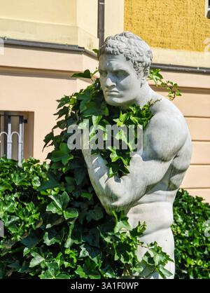 Eine Marmorstatue eines muskulösen Mannes, teilweise bedeckt mit grünem Efeu, stehend an einer gelben Wand. Die Statue hat einen ernsten Ausdruck und ist umgeben Stockfoto