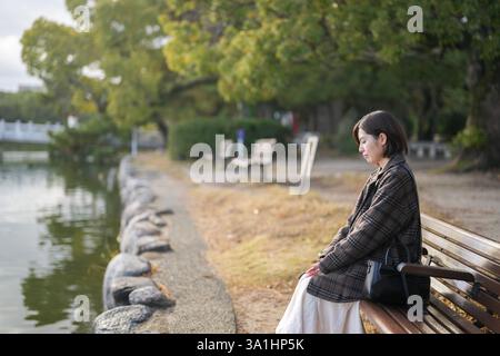 Winter im Ohori Park in Chuo Ward, Fukuoka City, Japan. Eine 29-jährige Japanerin in einem karierten Mantel. Sie sitzt auf einer Bank am Pon Stockfoto