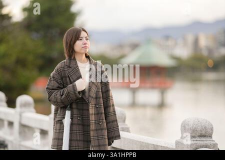 Winter im Ohori Park in Chuo Ward, Fukuoka City, Japan. Eine 29-jährige Japanerin in einem karierten Mantel. Sie geht über die Brücke des Teichs. Stockfoto