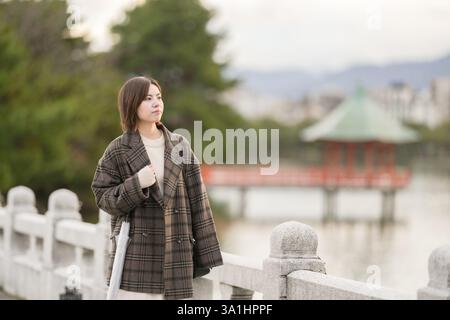 Winter im Ohori Park in Chuo Ward, Fukuoka City, Japan. Eine 29-jährige Japanerin in einem karierten Mantel. Sie geht über die Brücke des Teichs. Stockfoto