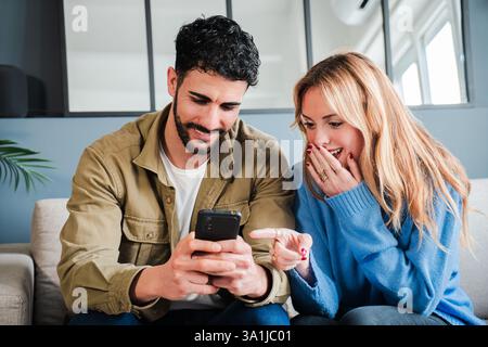 Ein aufgeregtes Paar entdeckt auf dem Smartphone eine Überraschung, während es zusammen auf einer gemütlichen Couch sitzt und einen wunderbaren Moment erzählt, der in einem herzerwärmenden Ausdruck der Verbindung festgehalten wird. Hochwertige Fotos Stockfoto