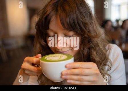Eine junge Frau mit lockigen Haaren genießt in einem warmen und einladenden Café den Duft eines Matcha Latte und genießt einen Moment der Ruhe in der lebhaften Atmosphäre Stockfoto