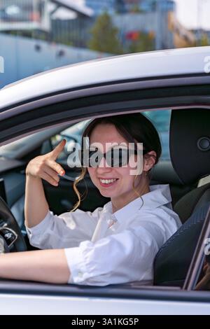 Lächelnde Frau in Sonnenbrille, die ein Auto fährt und eine spielerische Geste macht Stockfoto