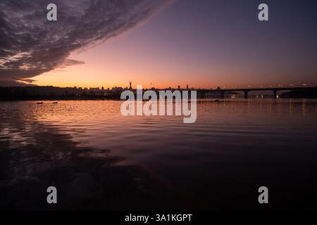 Während der Tag in die Nacht übergeht, färbt ein atemberaubender Sonnenuntergang den Himmel in lebendigen Tönen, erhellt einen ruhigen Fluss und beleuchtet die Skyline der Stadt, was eine bezaubernde Szene schafft, die Sie genießen können Stockfoto