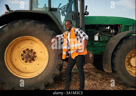 Lehnen Sie sich auf das große Rad des Traktors. Der schöne Afroamerikaner ist auf dem landwirtschaftlichen Gebiet. Stockfoto
