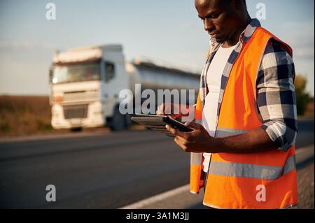 In orangefarbener Uniform, mit Tablette. Schwarzer Mann steht auf der Straße mit einem Truck drauf. Stockfoto