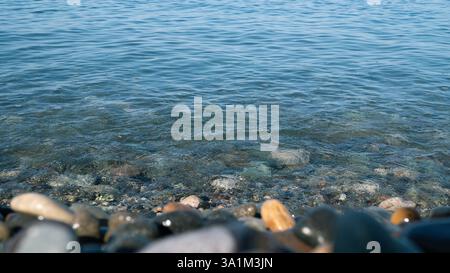 Eine ruhige Küstenlinie mit glatten, bunten Kieselsteinen im Vordergrund und klarem, kräuselndem blauem Wasser, das sanft über sie fließt. Sonnenlicht wird auf dem reflektiert Stockfoto