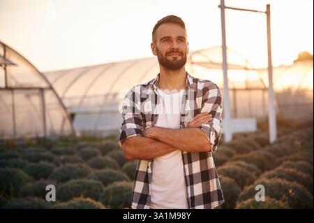 Ernstzunehmender junger Mann auf dem Ackerfeld steht mit gekreuzten Armen in der Nähe des dahinter liegenden Gewächshauses. Stockfoto
