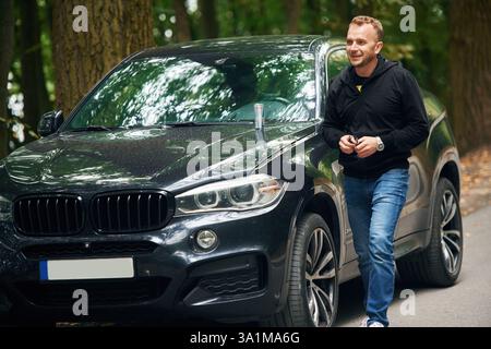 Ein Mann in Jeans ist draußen in der Nähe seines modernen schwarzen Autos. Stockfoto