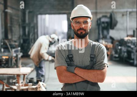 Stehend mit gekreuzten Armen. Junger Fabrikarbeiter in grauer Uniform. Stockfoto