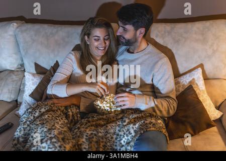 Ein romantisches Paar teilt sich eine Schüssel Popcorn, während es abends auf dem Sofa einen Film ansieht und einen gemütlichen und intimen Moment miteinander genießt Stockfoto