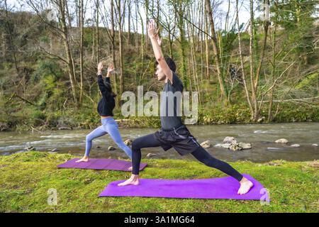 Junger Mann und Frau, die Yoga-Posen auf Matten am Fluss in einer friedlichen natürlichen Umgebung durchführen, in einem gesunden Lebensstil Stockfoto