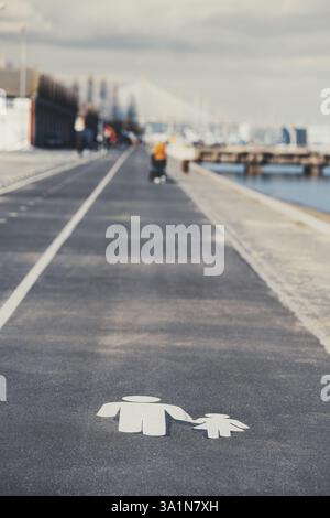 Fußgängerweg mit einem weiß bemalten Symbol eines Erwachsenen, der die Hand eines Kindes auf dem Asphalt hält. Der verschwommene Hintergrund zeigt eine Uferpromenade, einen Radfahrer Stockfoto