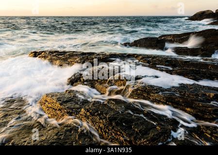 Wellen krachen an der schwedischen Küste gegen die Felsen und schaffen ein dynamisches Zusammenspiel von Wasser und Stein bei Sonnenuntergang. Das goldene Licht verstärkt die Schönheit Stockfoto