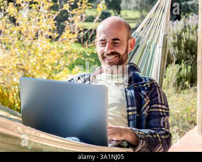 Lächelnder Mann, der in der Hängematte an einem Laptop arbeitet Stockfoto