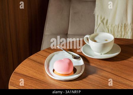 Herzförmiger Mousse-Kuchen mit rosa Velours und einer Tasse Latte auf einem Tisch in einem Café. Stockfoto