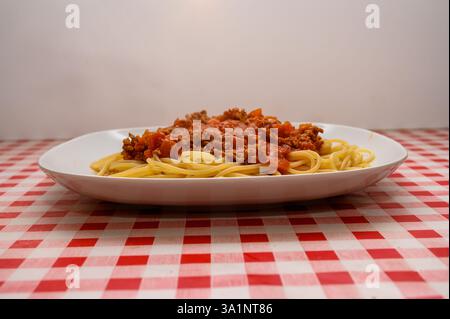 Eine großzügige Portion Spaghetti mit reichhaltiger Fleischsauce wird auf einem weißen Teller serviert. Die markante rot-weiße karierte Tischdecke verleiht dem Besucher einen besonderen Touch Stockfoto