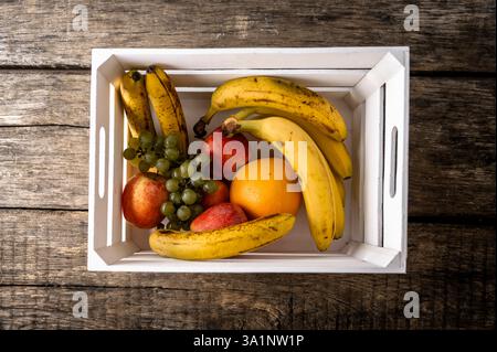 Verschiedene frische Früchte, darunter Bananen, Äpfel, Trauben und eine Orange, in einer weißen Holzkiste auf einem rustikalen Holztisch angeordnet. Stockfoto