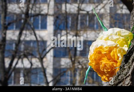 Bunte Stoffblumen zieren einen Baumstamm und bilden einen fröhlichen Kontrast zu den modernen Gebäuden im Hintergrund. Stockfoto