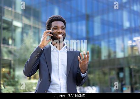 Lächelnder, freudiger Geschäftsmann, der vor dem Bürogebäude auf dem Handy spricht. Erfolgreicher afroamerikanischer Mann im Business-Anzug, der in der Stadt spaziert. Stockfoto