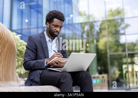 Ein fokussierter Geschäftsmann im Anzug arbeitet im Freien an einem Laptop, während er Notizen schreibt und Ohrstöpsel vor einem modernen Gebäude trägt. Stockfoto