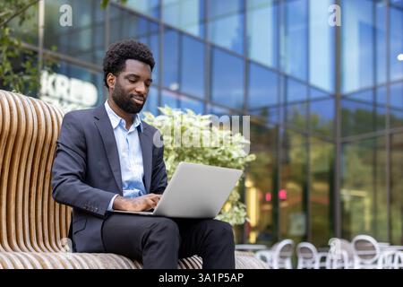 Ein junger Profi arbeitet ferngesteuert und nutzt seinen Laptop, während er auf einer modernen Bank mit einem Bürogebäude als Hintergrund sitzt. Stockfoto