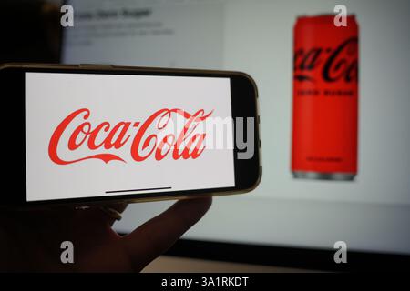 Roosendaal, Niederlande - 10. März 2025: Ein Mobiltelefon mit dem Logo von Coca-Cola, mit der Website des Unternehmens und dem Produkt im Hintergrund. Stockfoto