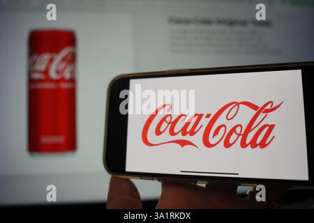 Roosendaal, Niederlande - 10. März 2025: Ein Mobiltelefon mit dem Logo von Coca-Cola, mit der Website des Unternehmens und dem Produkt im Hintergrund. Stockfoto