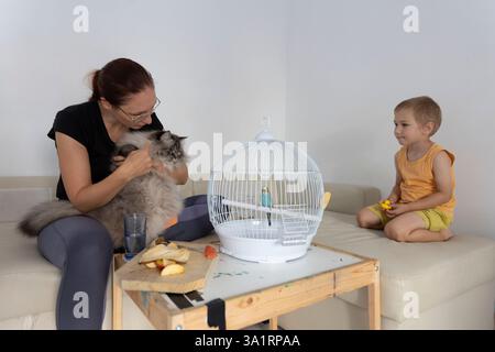 Mutter hält Katze, während der Junge Budgie zu Hause im Käfig beobachtet Stockfoto