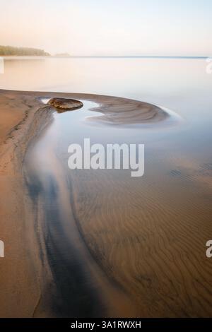 Das Licht am frühen Morgen offenbart das friedliche Wasser des Lake Vanern in Schweden, während sanfte Wellen das Sandufer streicheln und ein einsamer Felsen am Wasser steht Stockfoto