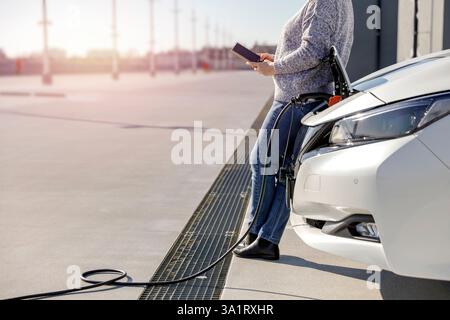 Eine Frau mit einem Smartphone neben einem weißen Elektroauto, das an ein Ladekabel auf einem gepflasterten Parkplatz angeschlossen ist. Stockfoto