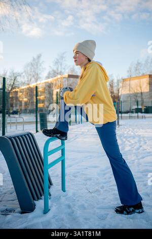 Junge Frau, die sich auf Outdoor-Fitnessgeräten in einem schneebedeckten Park ausdehnt Stockfoto
