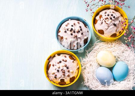 Köstliche Ostercupcakes und Eier auf hellblauem Hintergrund mit Blumen. Osterkuchen mit Glasur in hellen Backformen in Gelb- und Himmelsfarbe. Co Stockfoto