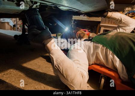 Fokussierter Mechaniker zur Inspektion und Reparatur des Fahrzeugs mit Taschenlampe unter dem Fahrzeug in der Werkstatt. Stockfoto