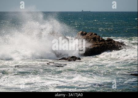 Wellen, die unter hellem Sonnenlicht auf dem offenen Meer gegen die felsige Küste krachen Stockfoto