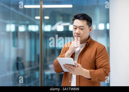 Ernsthaft denkender asiatischer Mann im Büro mit Tablet-Computer. Geschäftsmann in lässiger Kleidung, der neben dem Fenster steht. Stockfoto