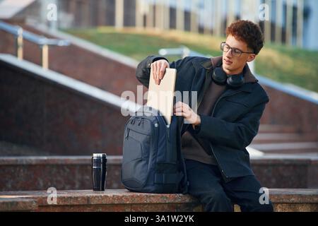 Nehmen Sie den Laptop aus dem Rucksack. Der junge Student ist außerhalb der Universität, in schwarzer Kleidung. Stockfoto