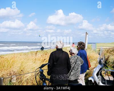 Egmond an Zee, Niederlande - 25. August 2024: Drei Frauen stehen in Egmond aan Zee, Niederlande mit Blick auf den Strand und das Meer. Ein Fahrrad und ein Roller parken in der Nähe, was ein Gefühl von Zeit und Freundschaft weckt. Stockfoto