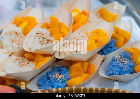 Traditionelles thailändisches Dessert Mango Klebreis in kleinen blattförmigen Tellern, mit weißem und blauem Klebreis, Sesamsamen und frischen reifen Mangowürfeln. Stockfoto