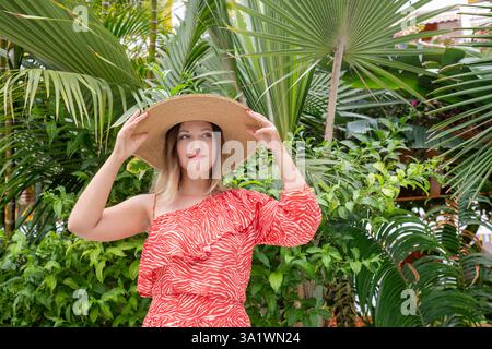 Stilvolle Frau mit einem auffälligen roten Einschulterkleid mit Zebramuster und breiter Strohhut in einem üppigen Garten Stockfoto