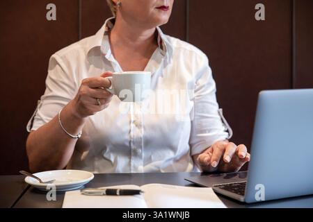 Eine Frau mittleren Alters genießt einen Kaffee, während sie an ihrem Laptop arbeitet, eine Frau, die eine Tasse Kaffee in der einen Hand hält und mit der anderen schreibt Stockfoto