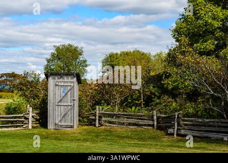 Rustikales öffentliches Outthouse am Rande eines Parks mit Zedernzäunen und blauem Wolkenhimmel. Stockfoto