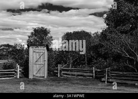 Rustikales Outthouse neben einem geteilten Eisenbahnzaun und Bäumen in Schwarz-weiß Stockfoto