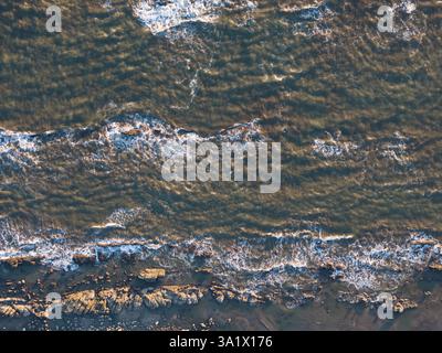 Sonnenlicht wirft goldenes Licht auf die Meereswellen, während sie bei Sonnenuntergang sanft gegen die felsige Küste krachen. Stockfoto