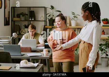 Große Aufnahme von afroamerikanischer Chefin in kluger Kleidung, die der neuen Programmiererin mit Down-Syndrom den Schreibtisch demonstriert, die ihre Dokumente hält Stockfoto