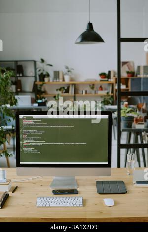 Vertikale Aufnahme eines silbernen Computers mit Codes auf dem Bildschirm auf einem Holztisch in einem modernen Büro, dekoriert mit grünen Pflanzen in Töpfen Stockfoto