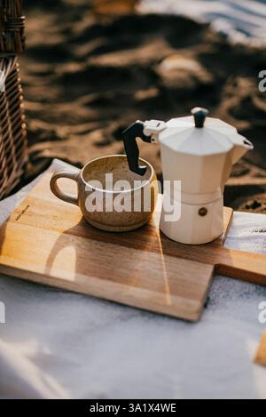 Eine Kaffeekanne und eine Tasse sind auf einem Holzbrett. Die Kaffeekanne ist weiß und die Tasse braun. Die Szene spielt an einem Strand, mit dem Sand und dem Boardprofi Stockfoto